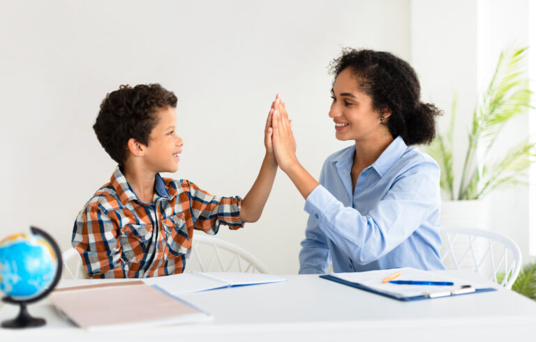 Successful School Day. Cheerful Schoolboy And Teacher Woman Giving High Five Celebrating Educational Success And Knowledge Progress, Sitting Studying At Table Indoors. Tutoring Concept